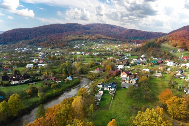 Luftaufnahme eines ländlichen Dorfes mit kleinen Häusern zwischen herbstlichen Berghügeln, die mit gelbem und grünem Fichtenwald bedeckt sind.