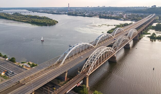 Luftaufnahme von Auto und Eisenbahn Darnitsky Brücke über Dnjepr Fluss. Skyline des Sonnenuntergangs der Stadt Kiew, Ukraine