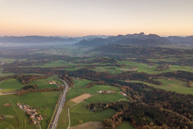 Luftaufnahme von kleinen verstreuten Bauernhäusern mit roten Ziegeldächern zwischen grünen Bauernfeldern und entferntem Bergwald im Sommer.