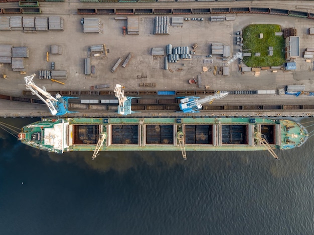 Luftaufnahme von oben am Dock mit Boot und Kränen. Hafenkräne laden verschiedene Ladungen oder Baumaterialien in den Laderaum des Schiffes. Warenlieferung auf dem Seeweg. Globale Wirtschaft.