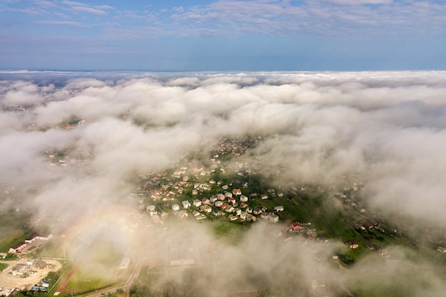 Luftaufnahme von weißen Wolken über einer Stadt oder einem Dorf mit Reihen von Gebäuden und kurvigen Straßen zwischen grünen Feldern im Sommer. Landschaft von oben.