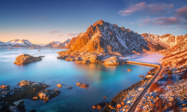 Luftaufnahme von wunderschönen Bergen, kleinen Inseln im Meer, blauer Himmel bei Sonnenuntergang auf den Lofoten-Inseln in Norwegen im Winter, Draufsicht auf die Straße, schneebedeckte Felsen, Steine, Meeresküste, klares Wasser, Draufsicht