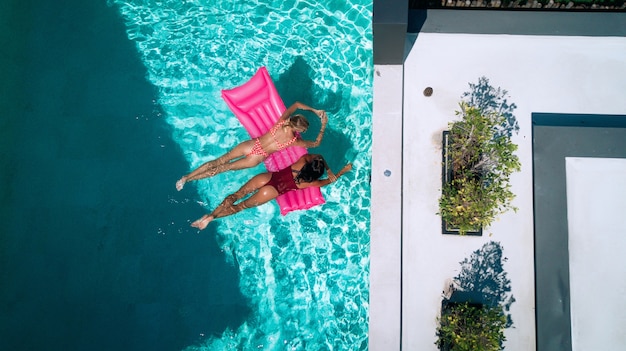 LUFTAUFNAHME Zwei fröhliche junge Frauen genießen auf aufblasbaren Schwimmern im Pool zu plaudern