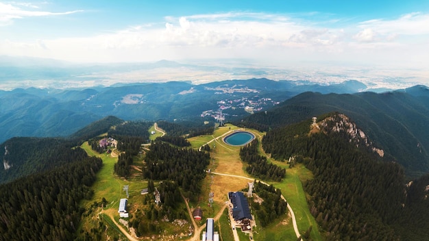 Luftdrohnen-Panoramablick auf den rumänischen See Poiana Brasov und andere Gebäude auf dem Gipfel des Berges, der in der Ferne mit üppigen Wäldern bedeckt ist