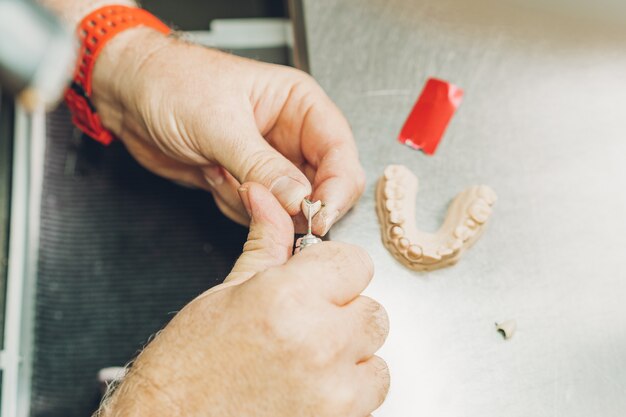 Luftnahaufnahme der Hände eines kaukasischen erwachsenen Mannes, die eine Zahnform mit einem elektrischen Mikromotor in einem auf dentale Knochenkeramik spezialisierten Dentallabor anpassen, um Porzellankronen herzustellen