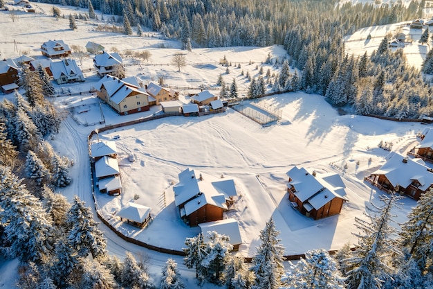 Luftwinterlandschaft mit kleinen Dorfhäusern zwischen schneebedecktem Wald in kalten Bergen am Abend.