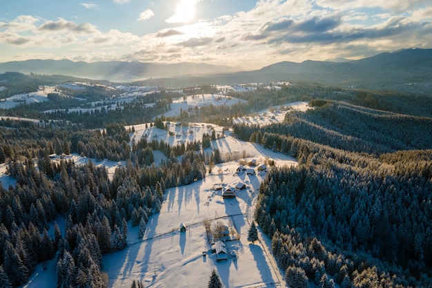 Luftwinterlandschaft mit kleinen Dorfhäusern zwischen schneebedecktem Wald in kalten Bergen am Abend.