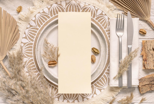 Lugar de mesa de casamento com cartão em branco vertical no prato na vista superior de placemat boêmio. Maquete do cartão de casamento Boho. Modelo bege tropical com plantas secas, folhas de palmeira e grama dos pampas, coloque texto para fotos