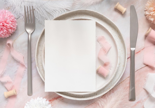 Lugar de la mesa de boda con una tarjeta en blanco cerca de flores, cintas de seda y plumas, vista superior. Plantilla romántica con tarjeta de papel vertical en un plato sobre una mesa de mármol plana. Maqueta de tarjeta, espacio de copia