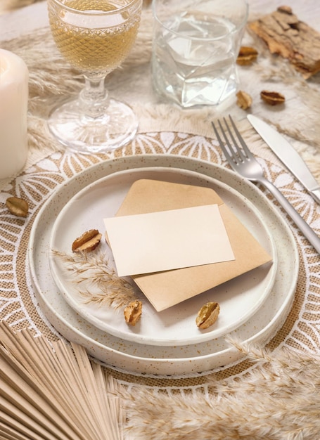 Lugar de mesa de boda con tarjeta en blanco y sobre en mantel bohemio de cerca. Maqueta de tarjeta de boda boho. Plantilla beige tropical con hojas de palmera secas y pasto de la pampa, colocar texto fot