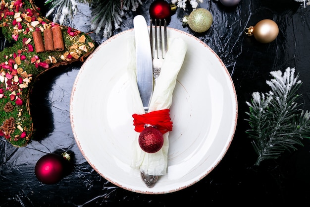 Lugar de mesa de Navidad. Plato blanco, cuchillo y tenedor con adornos navideños en mesa negra. Vista superior.