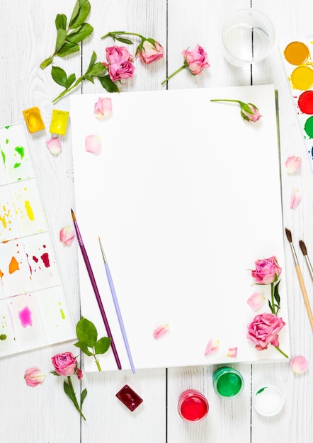 Lugar de trabajo del artista Pinceles de papel paleta de pinturas de acuarela y rosas sobre fondo blanco de madera Vista superior