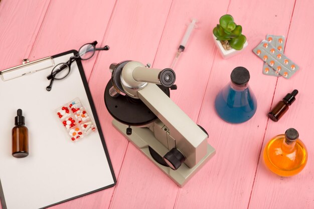 Lugar de trabajo científico médico microscopio píldoras jeringa anteojos matraces químicos con portapapeles líquido en mesa de madera rosa