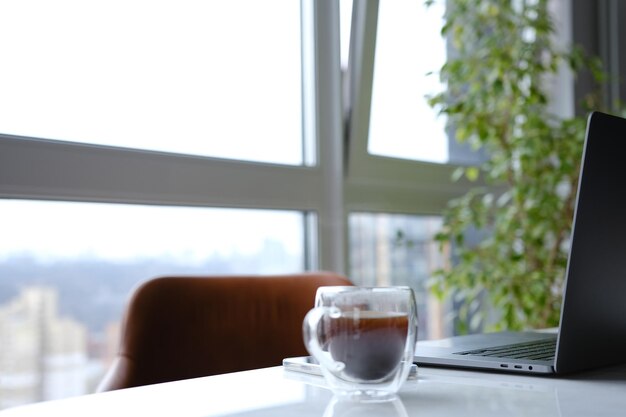 Foto lugar de trabajo con computadora portátil y una taza de café transparente en la oficina del hogar en un escritorio de mármol blanco con una silla de cuero marrón y un árbol verde en el fondo con una ventana