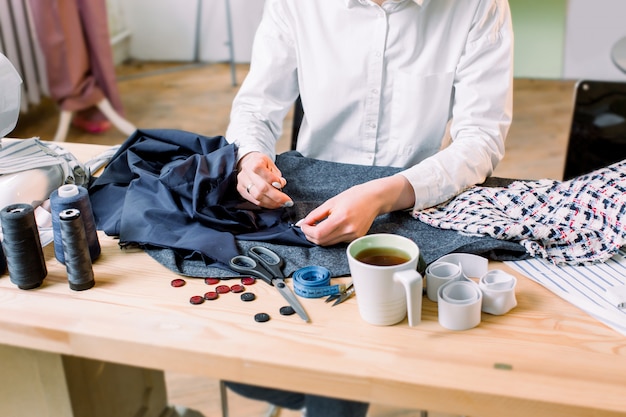 Lugar de trabajo de costurera. Recorte la imagen de las manos de la modista cosiendo un botón. Botones, materiales para pantalones, patrones, tijeras, hilos y agujas, cinta métrica y una taza de té sobre la mesa.