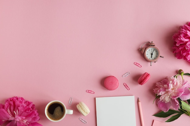 lugar de trabajo femenino con bloc de notas lápiz reloj despertador flores de peonía una taza de café y macarons