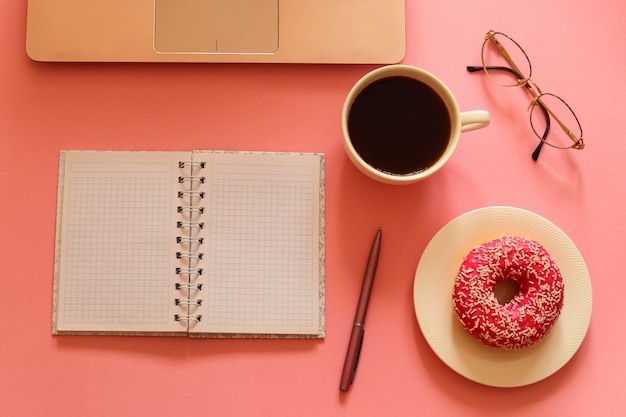 Lugar de trabajo femenino con laptop, donut, café, cuaderno, gafas y bolígrafo en la mesa rosa