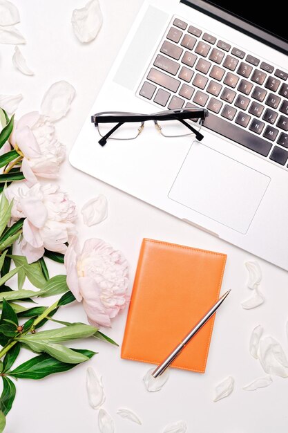 Lugar de trabajo con laptop, anteojos, cuaderno, pluma y flores de peonías en el fondo de la mesa blanca. Plano, vista superior