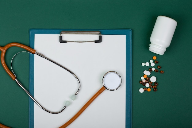 Lugar de trabajo médico - estetoscopio naranja, pastillas, botellas médicas y portapapeles vacío sobre fondo de papel verde