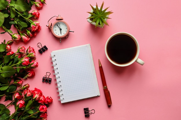 Lugar de trabajo de moda con flores de reloj despertador de bloc de notas y taza de café en rosa