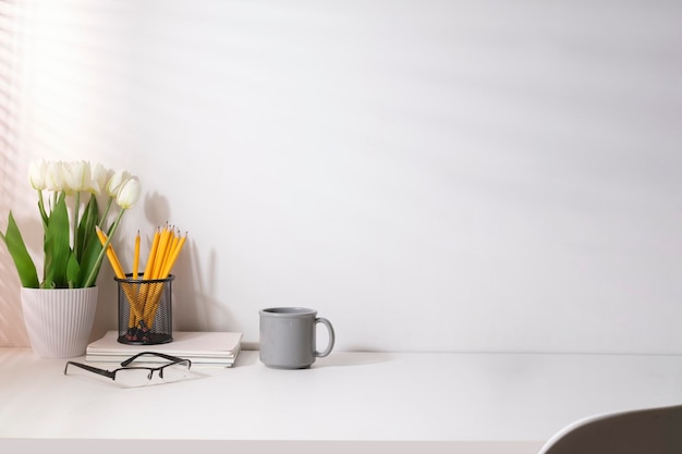 Lugar de trabajo moderno y femenino con maceta, taza de café y portalápices en mesa blanca. Copiar espacio para el texto publicitario.