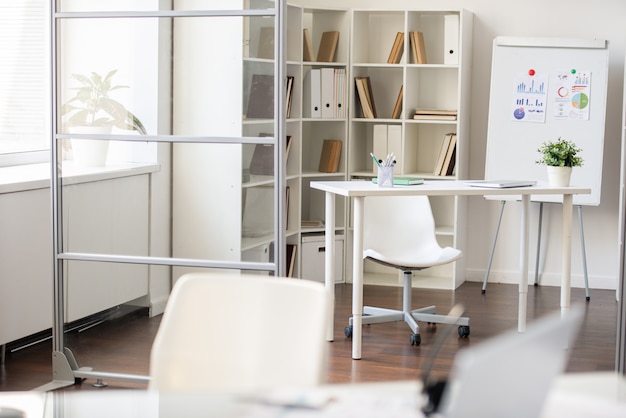 Lugar de trabajo de oficina moderno: mesa blanca y silla con ruedas colocadas cerca de estantes y pizarra con informes financieros