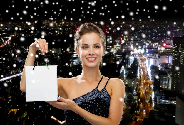 lujo, publicidad, vacaciones y concepto de venta - mujer sonriente con bolsa de compras en blanco sobre fondo de ciudad de noche nevada