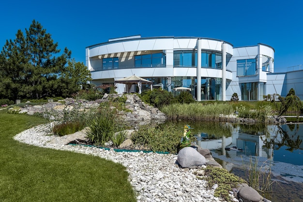 Lujosa y elegante casa de campo blanca de dos pisos con amplias ventanas y un patio trasero bien cuidado con un pequeño estanque artificial decorado con plantas ornamentales, guijarros y esculturas de jardín en verano