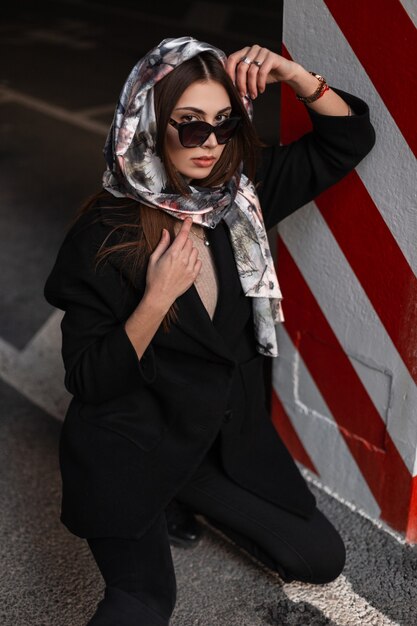 Lujosa mujer joven con gafas de sol con elegante pañuelo de seda en la cabeza con elegante abrigo negro está descansando sobre el asfalto cerca de la columna de rayas rojas y blancas en el estacionamiento. Modelo de moda atractiva chica de negocios.