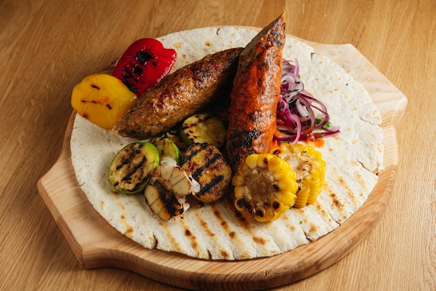 Lula Kebab mit Fladenbrot, Sauce, gegrilltem Mais und Zwiebeln auf einem Holzbrett