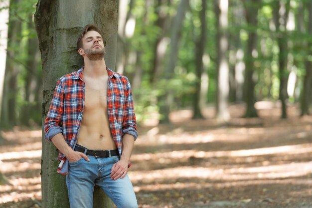 Lumbersexueller Kerl mit geschlossenen Augen im aufgeknöpften Holzfällerhemd-Waldhintergrund-Kopienraum