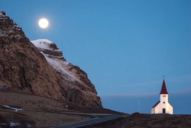 La luna llena se levanta sobre la iglesia de Vik en Islandia y una montaña con una capa de nieve