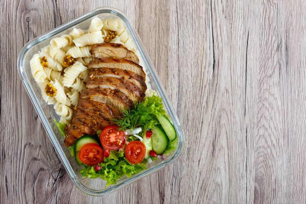 Lunchbox: Rotini mit gebackener Hähnchenbrust und Salat auf Holzhintergrund. Ansicht von oben, flach liegend. Leckeres gesundes Mittagessen. Das Konzept der gesunden Ernährung.