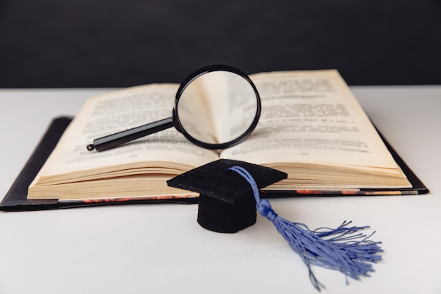 Lupa en libro abierto y gorro de graduación en mesa blanca. Concepto de educación.