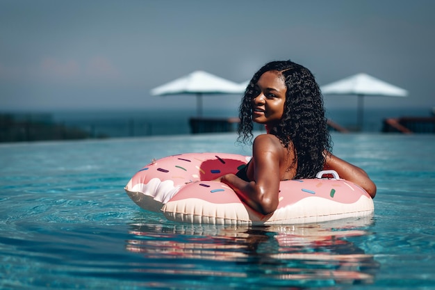 Lustige afroamerikanische Frau, die Spaß mit aufblasbarem Donut im Infinity-Pool hat; verschwommene Küstenlandschaft hinter ihr.