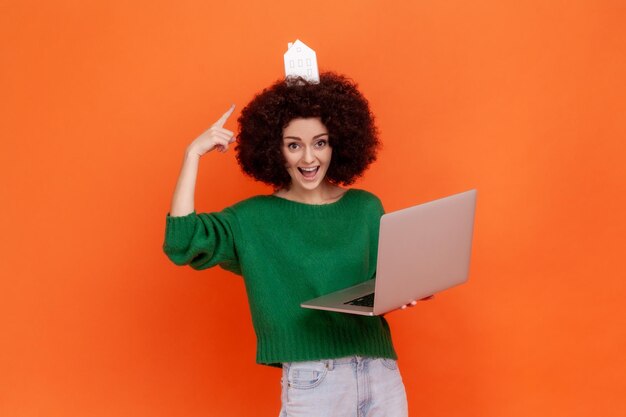 Lustige aufgeregte Frau mit Afro-Frisur, die einen grünen, lässigen Pullover trägt, der auf das Papierhaus auf dem Kopf zeigt und einen Laptop benutzt. Studioaufnahme im Innenbereich isoliert auf orangefarbenem Hintergrund.