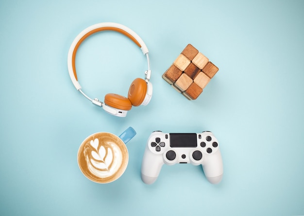 Lustige Dinge zu Hause: Gamepad, Kopfhörer, Holzpuzzlewürfel und Tasse Cappuccino auf blauem Hintergrund, Draufsicht. Ideenkonzept für Heimaktivitäten, minimaler flacher Lay-Stil von oben