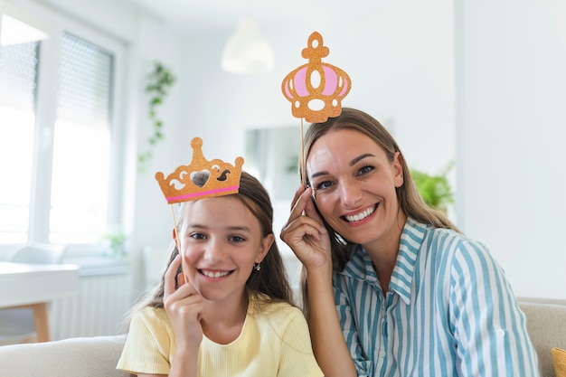 Lustige Familie auf einem Hintergrund der hellen Wand. Mutter und ihr Tochtermädchen mit Papierzubehör. Mutter und Kind halten Papierkrone auf Stock.