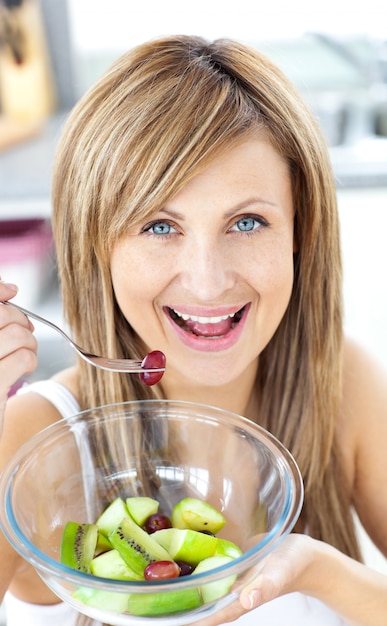 Lustige Frau, die einen Obstsalat lächelt an der Kamera in der Küche isst