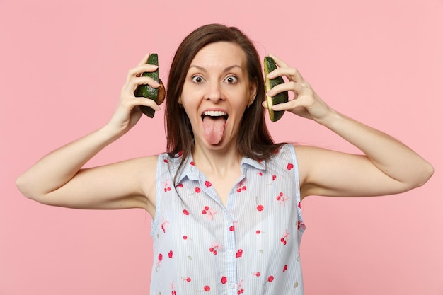 Lustige Frau in Sommerkleidung, die Zunge zeigt, die Hälften frischer, reifer Avocado-Früchte in der Nähe von Ohren hält, isoliert auf rosafarbenem Pastellhintergrund. Menschen lebendiger Lebensstil entspannen Urlaubskonzept. Mock-up-Kopienbereich.