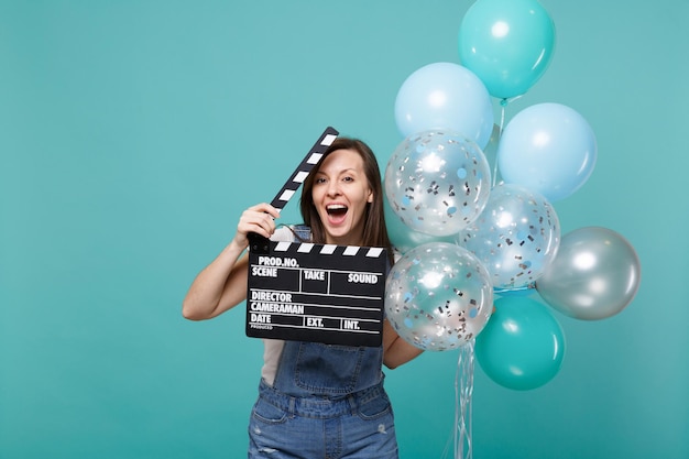 Lustige Frau mit offenem Mund halten klassischen schwarzen Film, der eine Klappe macht und mit bunten Luftballons feiert, die auf blauem türkisfarbenem Hintergrund isoliert sind. Geburtstagsfeier, Menschen Emotionen Konzept.