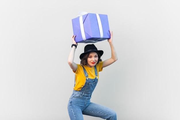 Lustige, fröhliche Frau in blauen Denim-Overalls, die eine große Geschenkbox über ihrem Kopf hält und in die Kamera blickt