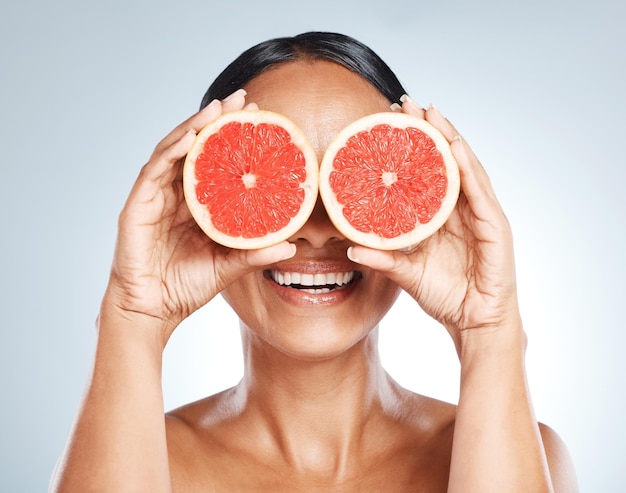 Lustige Frucht und Frau verwenden Grapefruit für einen Witz, der ihre Augen bedeckt, die vor einem blauen Studiohintergrund isoliert sind Gesundheitsschönheits- und Hautpflegemodell, das aufgrund von Vitamin C aus saftigen Zitrusfrüchten lächelt