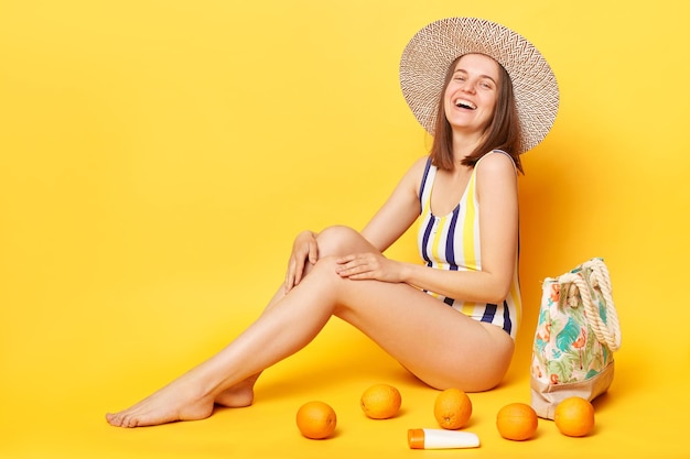 Lustige, glückliche Frau in gestreiftem Badeanzug und Hut, isoliert auf gelbem Hintergrund, sitzt lachend und hat eine gute Zeit im Sommerresort