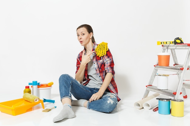 Lustige hübsche Frau in Freizeitkleidung, die mit gelben Handschuhen auf dem Boden sitzt, Instrumente für die Renovierungswohnung isoliert auf weißem Hintergrund. Tapetenzubehör für Malwerkzeuge. Hauskonzept reparieren