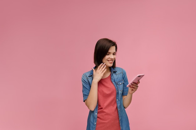 Foto lustige junge brünette, die rosa smartphone hält und lächelt