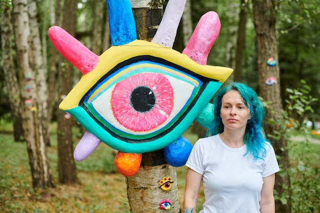 Lustige junge erwachsene Frau mit gefärbten türkisfarbenen Haaren in weißem T-Shirt in der Nähe einer Big-Eye-Kunstinstallation