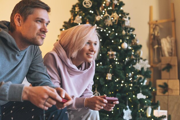 Lustige junge Familie in Heimkleidung, die Videospiele auf der Couch vor dem Hintergrund des Weihnachtsbaums spielt