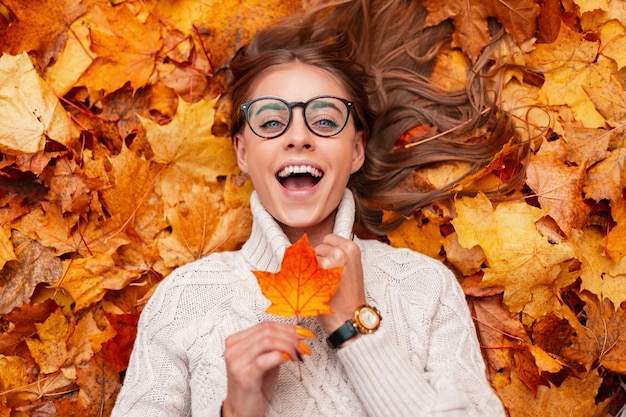 Lustige junge Hipster-Frau in einem modischen Strickpullover in stilvollen Gläsern mit einem orangefarbenen Blatt in den Händen drückt Überraschung aus, während sie auf Herbstlaub im Wald liegt. Positives Mädchenmodell, das sich ausruht.