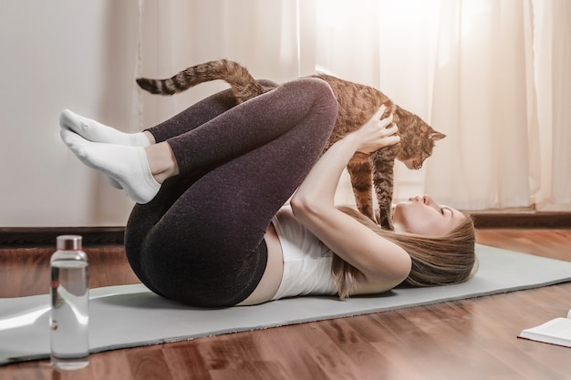 Lustige junge kaukasische Frau praktiziert Yoga zu Hause mit einer Katze.